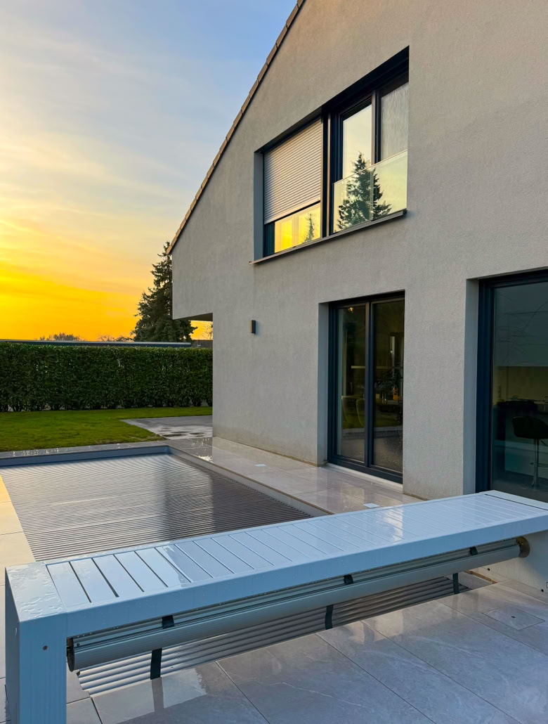 Modernes Haus mit großen Fenstern, Terrasse und Pool im Vordergrund, umgeben von einem gepflegten Garten bei Sonnenuntergang.
