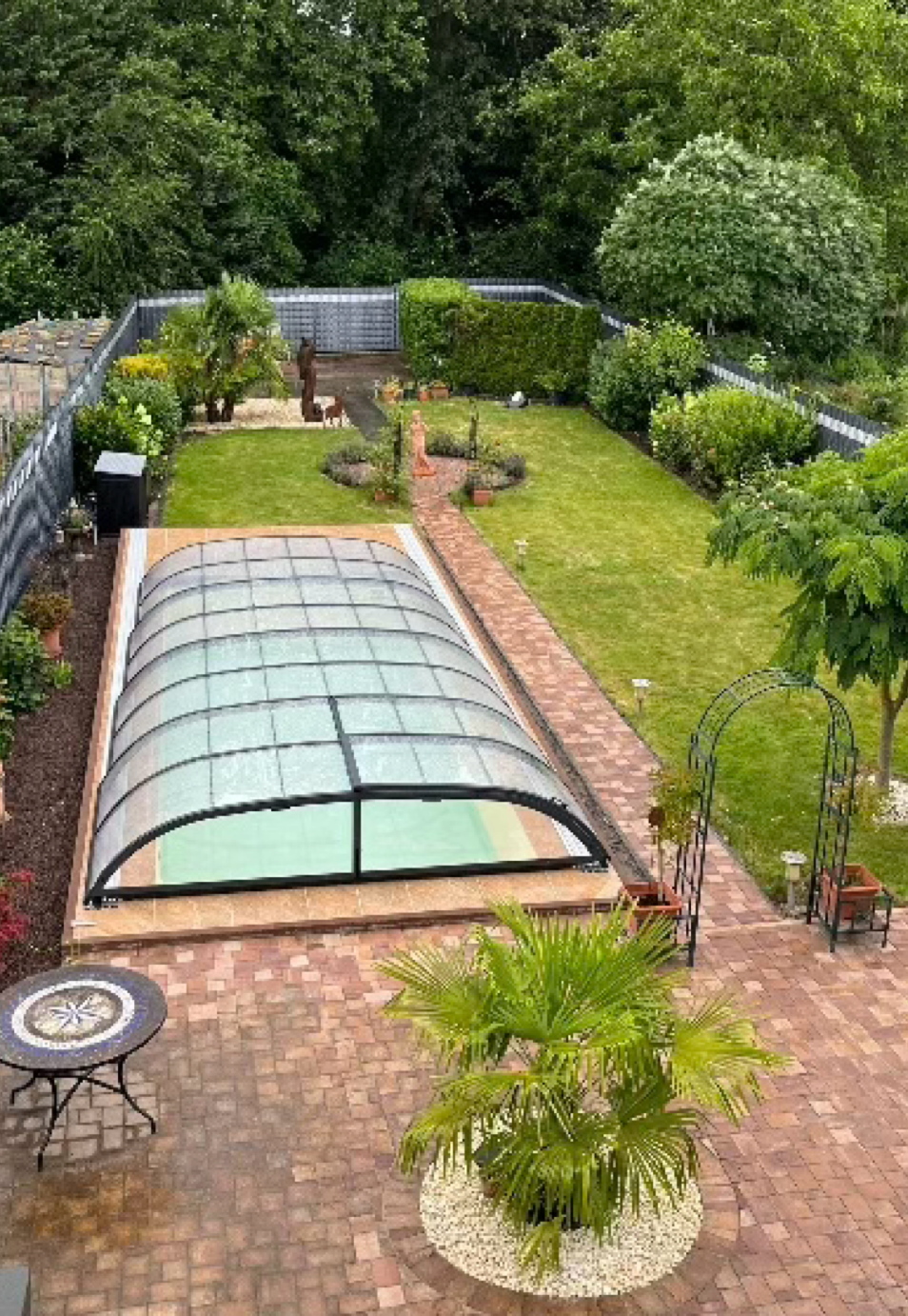 Garten mit überdachtem Pool, umgeben von Rasenflächen, Bäumen und Pflanzen; im Vordergrund ein runder Tisch mit Stühlen.